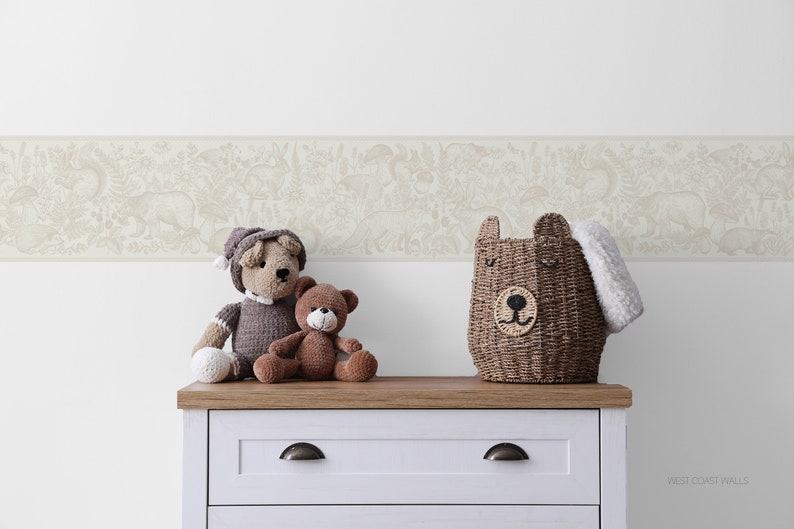 May include: A white dresser with a brown top and two drawers. The dresser has two silver knobs. On top of the dresser are three stuffed animals: a brown bear, a grey bear, and a brown and grey bear. There is also a brown wicker basket with a bear face on the front. The wall behind the dresser has a beige wallpaper border with a white background and a pattern of animals and plants.