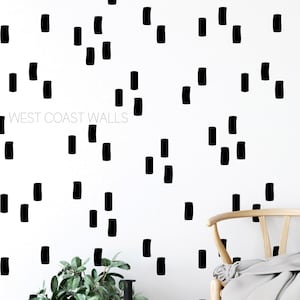 Peut inclure: Mur blanc avec un motif répétitif de formes rectangulaires noires. Le texte "WEST COAST WALLS" est visible. Un panier tressé avec une plante, un vase noir et une chaise en bois avec une couverture sont au premier plan.