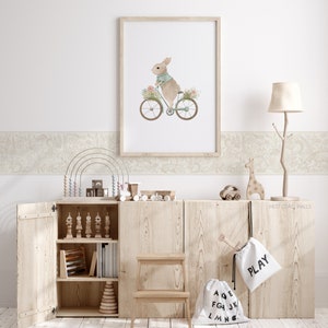 May include: A light wood cabinet with open doors revealing a variety of children's toys, including a wooden abacus, stacking rings, and a wooden giraffe. There is a white canvas bag hanging on the cabinet with the word "PLAY" printed on it. A framed print of a rabbit riding a bicycle with flowers is hanging on the wall above the cabinet.