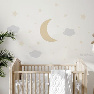 Könnte beinhalten: Ein Holzbettchen mit weißer Steppdecke und Bettwäsche. Die Wand dahinter zeigt ein Design aus Mond, Sternen und Wolken in sanften Beige- und Grautönen. Die Worte "WEST COAST WALLS" sind auf der Wand aufgedruckt.