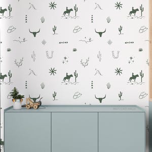 May include: White wallpaper featuring a repeating pattern of cowboy-themed illustrations in dark green. The design includes cowboys on horses, cacti, hats, bull skulls, and other desert-inspired elements. The wallpaper is displayed above a light blue cabinet.