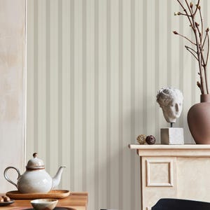 May include: A white ceramic teapot with a lid and a spout, sitting on a wooden table with a wooden plate and a small brown bowl. The background is a wall with vertical stripes in beige and white.