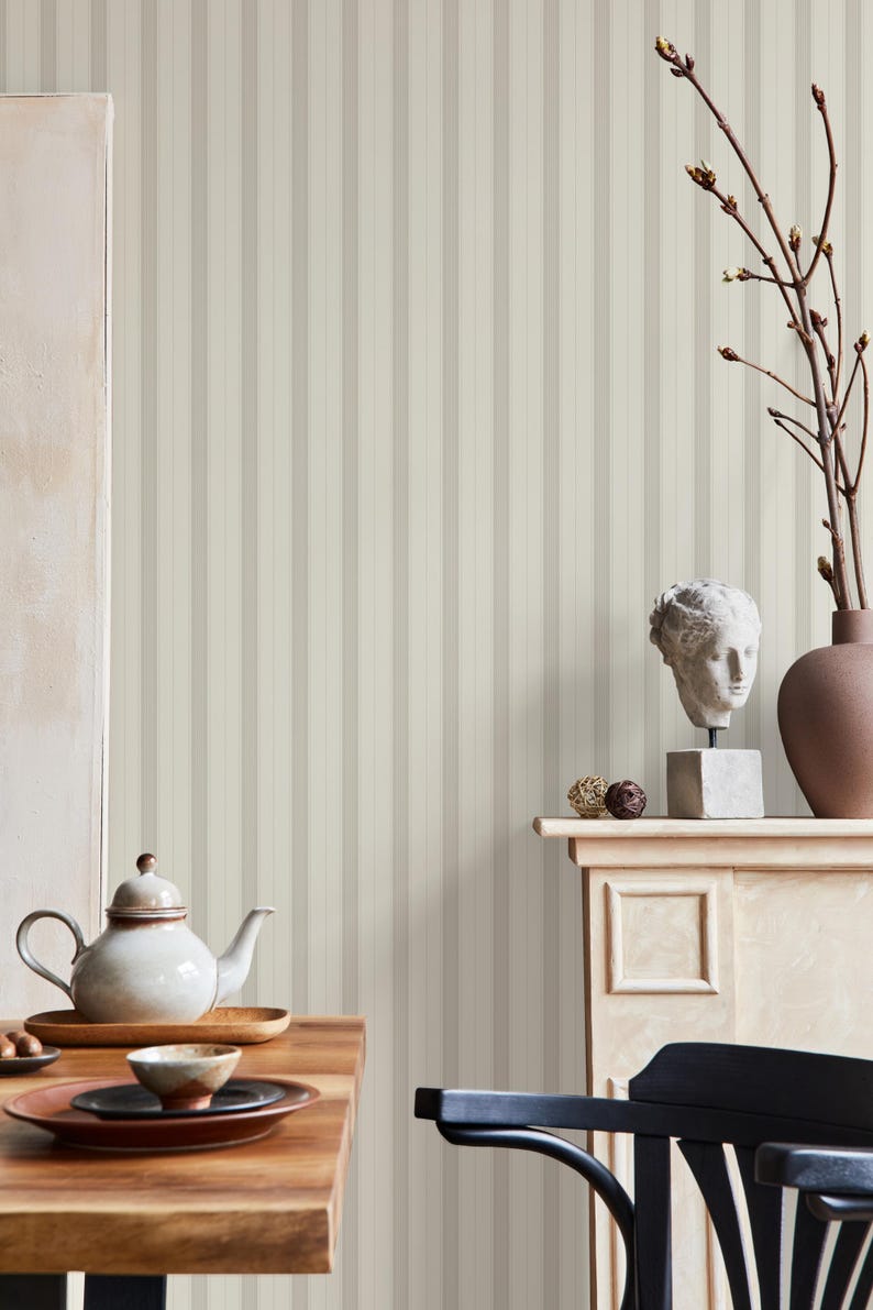 May include: A white ceramic teapot with a lid and a spout, sitting on a wooden table with a wooden plate and a small brown bowl. The background is a wall with vertical stripes in beige and white.
