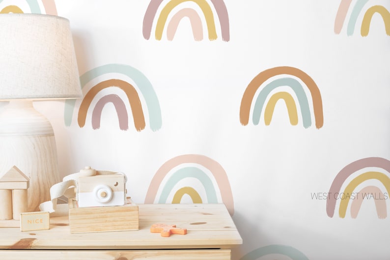 May include: A white wall with a repeating pattern of colorful rainbow decals. The rainbows are painted in shades of pink, blue, yellow, and brown. The wall is decorated with a wooden lamp, a wooden camera, and a wooden block that says "NICE".