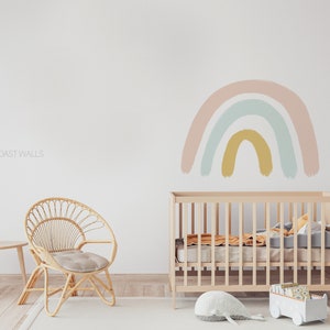 May include: A light pink, blue, and yellow rainbow wall decal above a wooden crib in a nursery. The crib has a white mattress and a white stuffed whale on the floor in front of it.