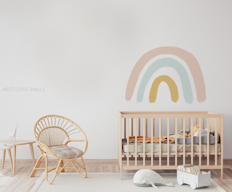 May include: A light pink, blue, and yellow rainbow wall decal above a wooden crib in a nursery. The crib has a white mattress and a white stuffed whale on the floor in front of it.