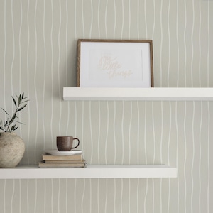 May include: Two white floating shelves against a light green wall with vertical white wavy lines. The top shelf holds a framed print with gold text. The bottom shelf displays a vase with greenery, a mug, and books.
