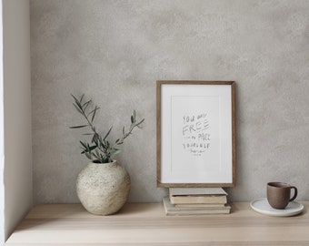 Può includere: Una stampa incorniciata con il testo "You are FREE to place yourself here." La stampa è su uno scaffale di legno con un vaso di verde, una pila di libri e una tazza da caffè marrone su un piattino bianco.