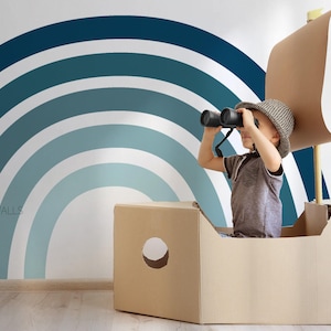 May include: A child wearing a hat looks through binoculars while sitting in a cardboard box boat. A blue and white rainbow wall decal is in the background. The cardboard boat has a cardboard sail.