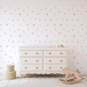 Op de afbeelding: Een witte commode met zes lades en roségouden knoppen staat voor een muur met een roze strikpatroon. Een beige schommelpaard en dozen staan op een wit tapijt.