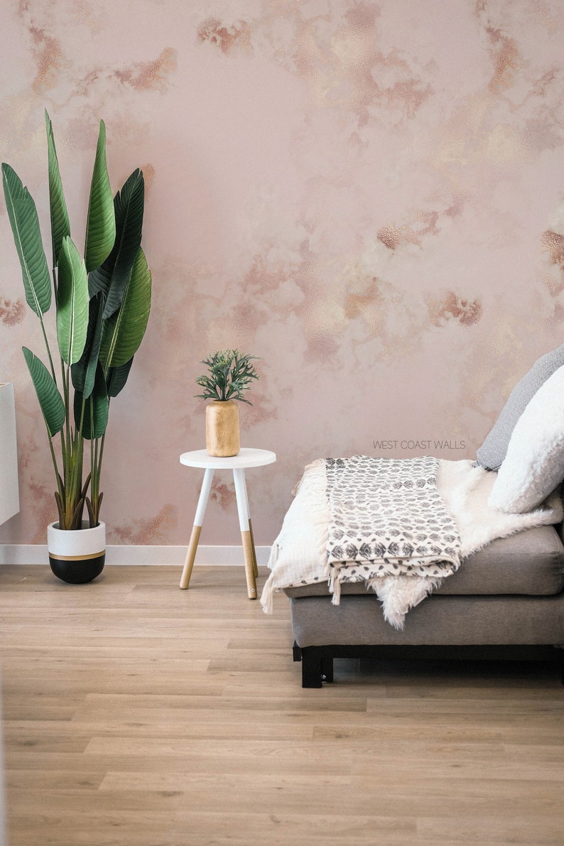 Può includere: Un muro rosa chiaro con un effetto acquerello, che presenta un tavolino bianco con un vaso dorato e una pianta verde. Un divano grigio con una coperta bianca e cuscini è in primo piano. Il testo 'WEST COAST WALLS' è visibile sul muro.