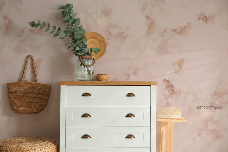Può includere: Una cassettiera bianca con quattro cassetti e maniglie in ottone è posizionata contro un muro rosa con un sottile motivo marmoreo. Una borsa di tela intrecciata è appesa al muro, e un vaso con rami di eucalipto e un vassoio rotondo in legno con una piccola ciotola sono sopra la cassettiera. Il testo 'WEST COAST WALLS' è visibile nell'angolo in basso a destra dell'immagine.