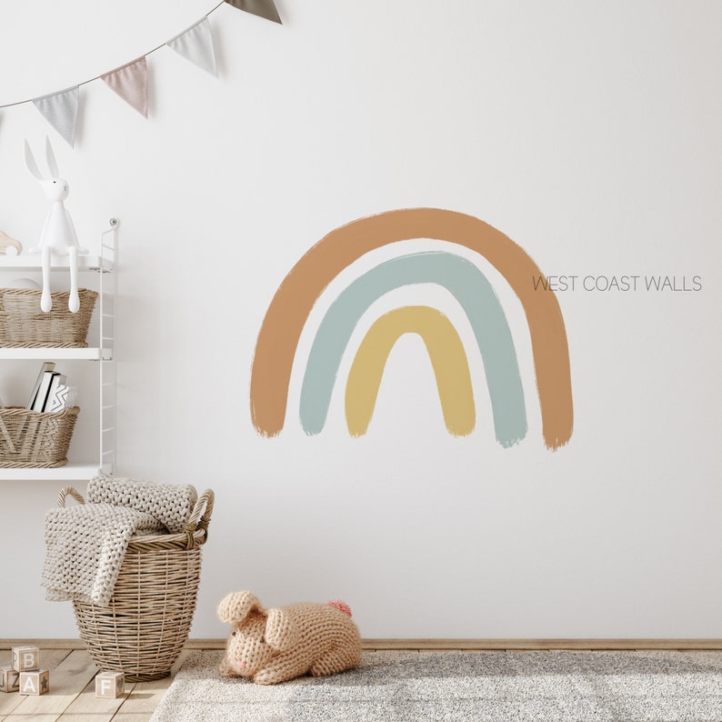 May include: A colorful rainbow wall decal with a light brown, light blue, and yellow color scheme. The decal is on a white wall in a nursery room with a white shelf, a basket, and a stuffed bunny.
