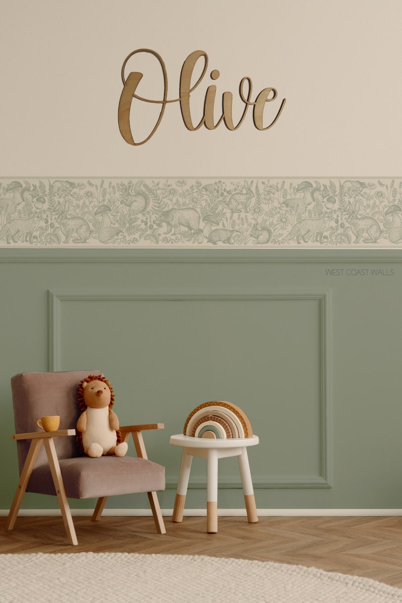 May include: A light green wall with a white border and a wooden name sign that says "Olive". The wall has a wallpaper border with a woodland animal design. A brown chair with a stuffed hedgehog sits next to a white stool with a rainbow wooden toy on top.