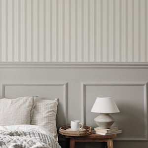 May include: A white bed with a gray and white striped headboard and a gray and white patterned blanket. There is a white ceramic mug on a wooden tray next to a lamp with a white shade on a wooden table.