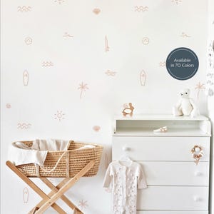 Può includere: Una scena di asilo nido con una parete bianca decorata con decalcomanie a tema spiaggia marrone chiaro. Una culla in vimini su un supporto in legno, un comò bianco e una tutina bianca con pompon sono visibili. Il testo "Available in 70 Colors" è in un cerchio.