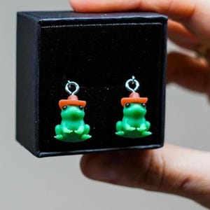 May include: A pair of green frog earrings, each wearing a small orange hat. The earrings are displayed in a black box. The frogs have detailed features and are suspended from silver-colored hooks.