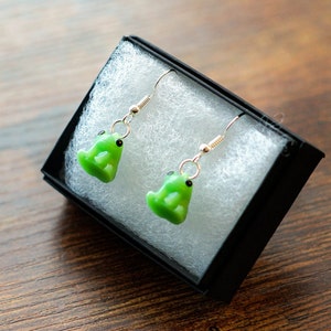 May include: A pair of green frog dangle earrings with silver hooks. The earrings are displayed in a black box with white padding.