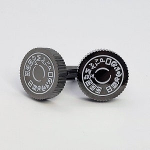 May include: A pair of black cufflinks with a silver camera aperture setting design. The cufflinks feature the numbers 1, 2, 4, 8, 16, 32, 64, 125, 250, 500, 1000, 2000, 4000, and the letters A, M, P, Z, and the words "A-DEP" and "M-DEP".