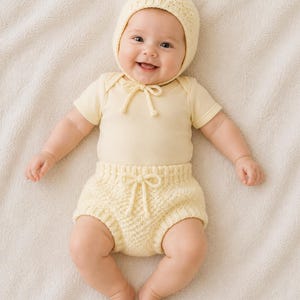 Könnte beinhalten: Ein lächelndes Baby trägt eine hellgelbe Strickmütze, einen kurzärmeligen Body und eine passende Windel. Das Outfit hat eine Schleife am Hals und in der Taille. Das Baby liegt auf einer cremefarbenen Decke.