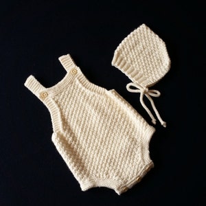 May include: A white knitted baby romper and matching bonnet. The romper has a button closure and the bonnet has a tie closure.