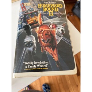 May include: A VHS tape of the Disney movie "Homeward Bound II: Lost in San Francisco." The cover features a golden retriever, a white dog, and a cat running on train tracks. The title is in yellow and red, with the text "Totally Irresistible... A Family Winner!"