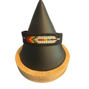Pode incluir: Uma pulseira de contas tecidas com fundo preto e um padrão horizontal central de contas brancas, vermelhas, laranja, amarelas e azuis. O design apresenta um motivo semelhante a uma pena criado com pequenas contas de vidro uniformes.