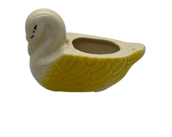 May include: A small ceramic planter shaped like a duck. The duck is white with yellow feathers and a small black eye. The planter has a small opening on the top for planting.