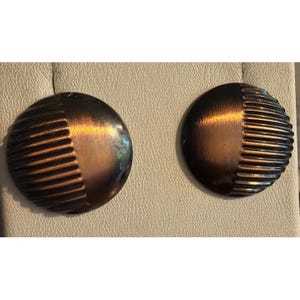 May include: A pair of circular stud earrings featuring a two-tone design. One half of each earring has a smooth, dark bronze-colored finish, while the other half displays a series of raised, vertical ridges with a metallic sheen.