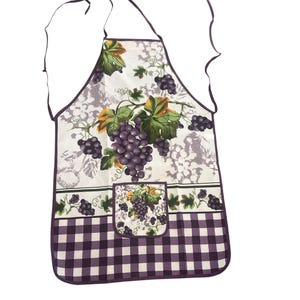May include: A white apron with a purple grape design and a purple and white checkered bottom. The apron features a pocket with a grape cluster design and green leaves. The apron has a purple trim and straps.