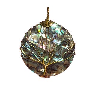 Puede incluir: Un colgante circular con un mosaico de piezas de concha de abulón iridiscente en tonos azules, verdes y morados. Un alambre de tono dorado está envuelto alrededor de la concha formando la silueta de un árbol con ramas sobre la superficie.
