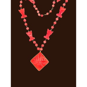 Peut inclure: Un collier en corail rouge avec un grand pendentif en corail rouge sculpté. Le pendentif est en forme de diamant et présente un motif géométrique. Le collier est composé de petites perles rondes en corail rouge.