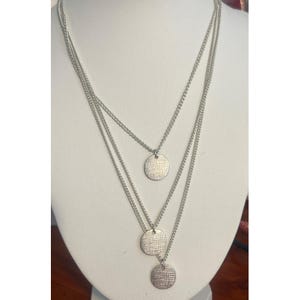 Puede incluir: Un collar plateado de tres hilos con colgantes circulares texturizados. Los colgantes cuelgan de delicadas cadenas, creando un efecto de capas. El collar se exhibe sobre un busto blanco, mostrando su diseño.