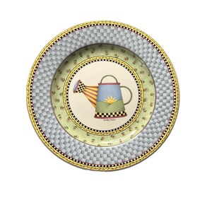 May include: A round ceramic plate with a chequered pattern in blue and grey. The centre of the plate features a colourful illustration of a watering can with a yellow spout and a green handle. The watering can is surrounded by a green wreath and pink flowers. The text "#Cherie Plum" is printed on the bottom of the plate.