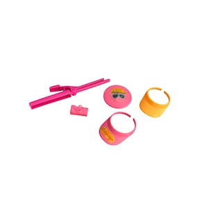 May include: A collection of pink and orange accessories, including a curling iron, a toy camera, a sun visor with "California" text, and a round item with sunglasses graphic. The items are arranged on a white surface.