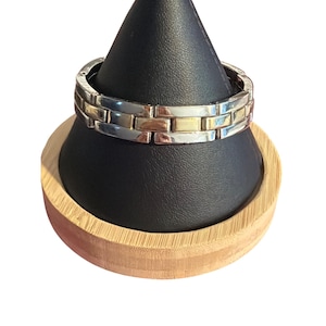 Könnte beinhalten: Ein silber- und goldfarbenes Armband mit einem ziegelartigen Design. Das Armband wird auf einem schwarzen, kegelförmigen Ständer präsentiert, der auf einem runden Holzsockel steht. Das Armband besteht aus rechteckigen Gliedern.