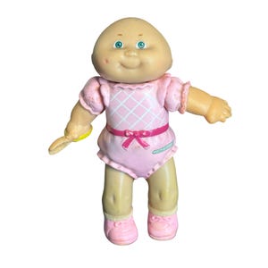 Könnte beinhalten: Eine kleine Cabbage Patch Kids Puppe mit Glatze, grünen Augen und heller Haut. Die Puppe trägt einen rosa Strampler mit weißem Rautenmuster am Oberteil, einen rosa Taillengürtel mit Schleife und rosa Schuhe. In einer Hand wird ein gelbes Zubehör gehalten.
