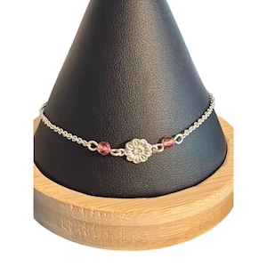 Op de afbeelding: Een zilveren armband met een bloemvormig middelpunt en roze kralen. De armband wordt gepresenteerd op een zwarte, kegelvormige standaard met een houten basis. De bloem heeft een klein kristal in het midden.