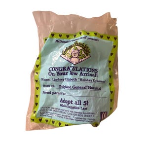 Può includere: Un giocattolo Happy Meal Cabbage Patch Kids di McDonald's. Il giocattolo è una bambola Cabbage Patch Kid con un vestito rosa e un cappello bianco. La confezione è blu e bianca con il testo "Congratulazioni per il tuo nuovo arrivo!" e "Adotta tutti e 5! Fino a esaurimento scorte."