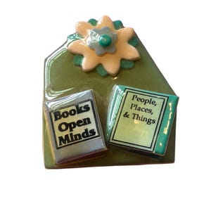 May include: A ceramic book-themed pin with a green background and a flower on top. Two small books are featured, one with the text "Books Open Minds" and the other with "People, Places, & Things."