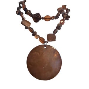 May include: A necklace featuring a large, circular, smooth brown pendant suspended from a strand of irregularly shaped, polished brown beads of varying sizes. The beads are strung in a double-layered arrangement, creating a cohesive brown color palette throughout the entire piece of jewelry.