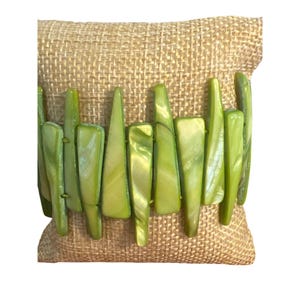 Puede incluir: Una pulsera de cuentas verdes con una mezcla de formas rectangulares y puntiagudas. La pulsera está hecha de cuentas verde claro con un brillo nacarado. La pulsera se exhibe sobre una superficie de arpillera beige.