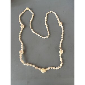 Puede incluir: Un collar hecho de pequeñas conchas de color crema. Las conchas están ensartadas, con conchas más grandes intercaladas en el diseño. El collar está sobre una superficie gris.