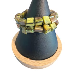May include: A two-strand bracelet with square, olive-green beads. The beads have a marbled appearance with brown and yellow tones. The bracelet is displayed on a black jewelry stand with a wooden base. The background is white.