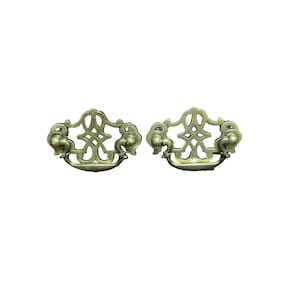 Może przedstawiać: Dwa antyczne mosiężne uchwyty do szuflad z ozdobnymi wzorami. Każdy uchwyt ma ozdobny, ażurowy środek i dwa okrągłe pokrętła po bokach.