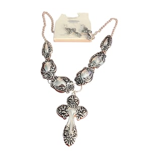 May include: A silver-coloured necklace and matching earrings set. The necklace has ornate oval links and a cross pendant. The earrings are small crosses. The jewellery is displayed on a white card.
