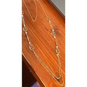 May include: A long gold-tone necklace with a delicate chain and alternating circular and rectangular links. The necklace features small, round, white pearls. The necklace is displayed on a wooden surface.