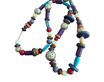 May include: A colorful beaded necklace with a variety of shapes and sizes. The necklace features beads in shades of purple, orange, turquoise, red, and gold. The beads are strung together with a gold-colored clasp.