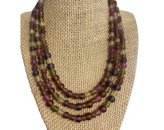 May include: A multi-strand necklace with a variety of earth-toned beads. The necklace features beads in shades of brown, green, and purple. The necklace is displayed on a burlap-covered bust.