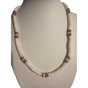 Puede incluir: Un collar de conchas blancas con detalles rosas y plateados. El collar presenta pequeñas cuentas de concha blancas y planas ensartadas, intercaladas con cuentas rosas de forma ovalada y espaciadores metálicos plateados.